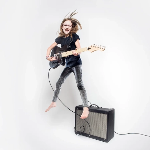 An elementary aged girl plays a black electric guitar on a white studio background. She laughs with joy as she jumps up in the air while rocking out.